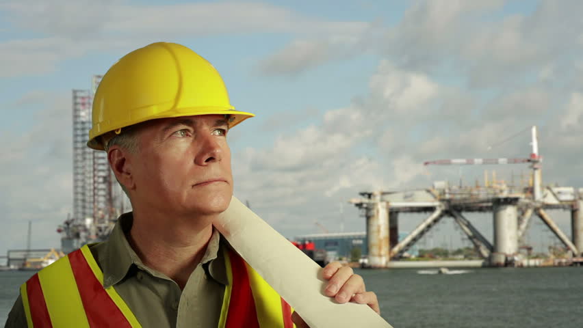 Drilling platforms are background for smiling man in hardhat and safety vest holding rolled up schematics or plans.