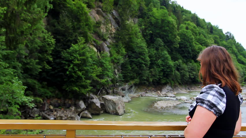 nice woman stands near railing bridge, then looks on waterfall in Carpathian Mountains