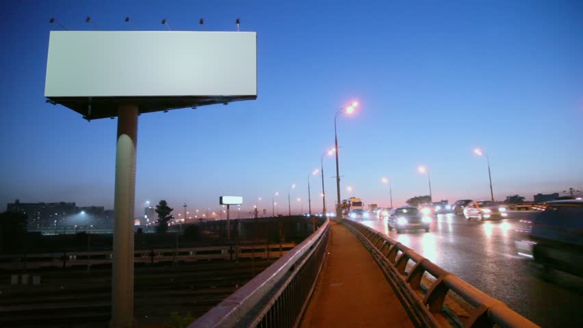 Empty advertising billboard on sidelines of road with traffic near railroad at evening, fast motion time lapse