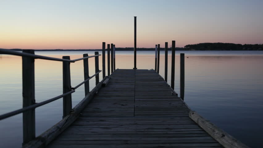 Sunset Dock - A weathered wooden dock with metal railings extends out into a gentry rippled sunset lake and a duck floats in the distance.