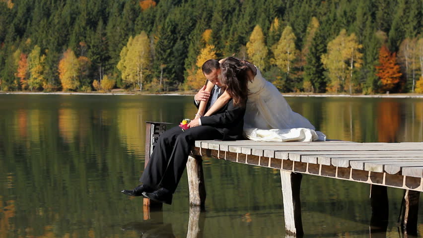 Beautiful young bride caressing her groom on the pontoon of a mountain lake reflecting an autumn colorful scenery in background