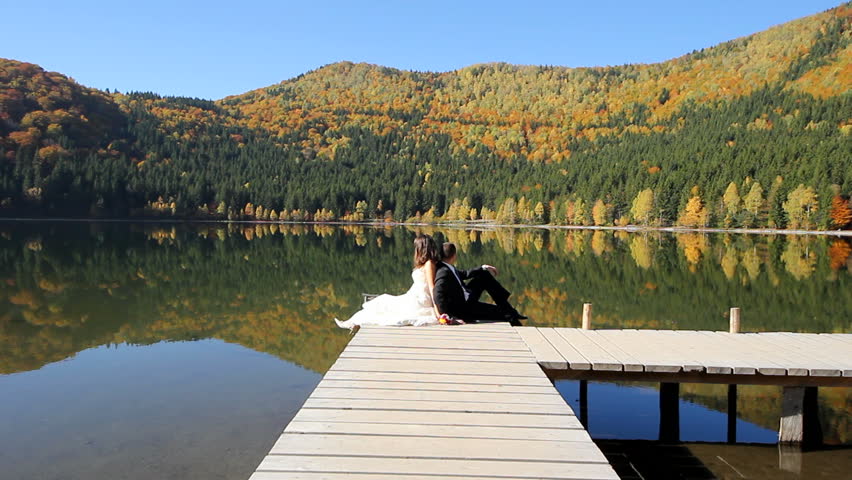 Bride and groom back to back sitting on a wooden pontoon on the shore of a lake reflecting a beautiful colorful autumn scenery