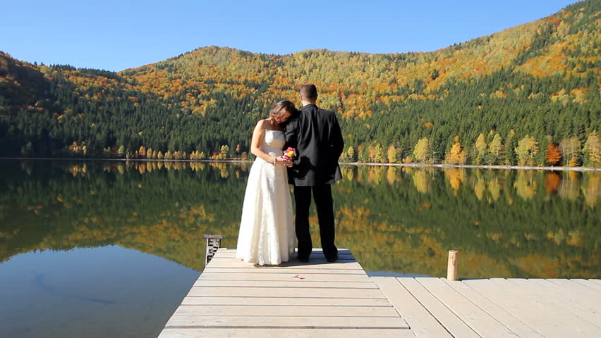 Bride head on groom's shoulder, both on a dock of a mountain lake, with a superb scenery in background