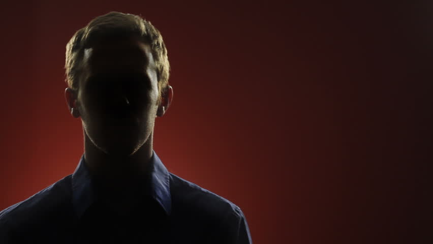 A businessman, whose face is hidden in shadow, stares into the camera in front of a red background. Centered version also available.