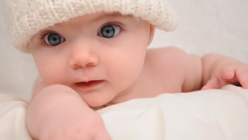 A cute little baby is looking into the camera and is wearing a white hat. The baby looks around and then smiles and laughs.