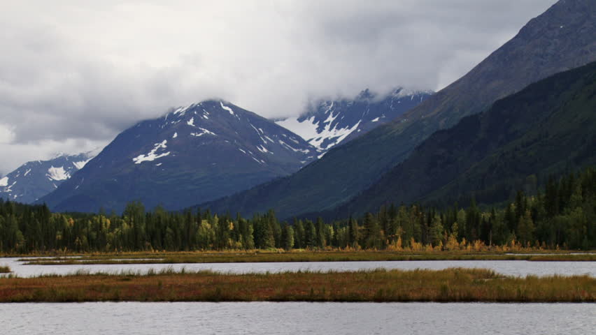 Shifting sunlight on an otherwise cloudy day briefly illuminates autumnal foliage on the shores of Tern Lake in Alaska, with solemn mountains beyond.