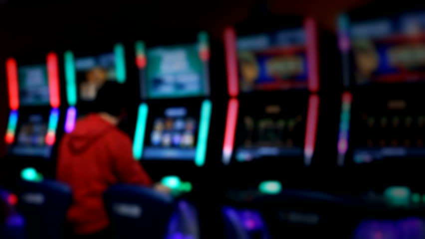 Slot machines videopoker man playing - A slot machine - video poker room, with lights glowing in the dark. A man, dressed in red, is playing at one machine. Bokeh - defocused shot.