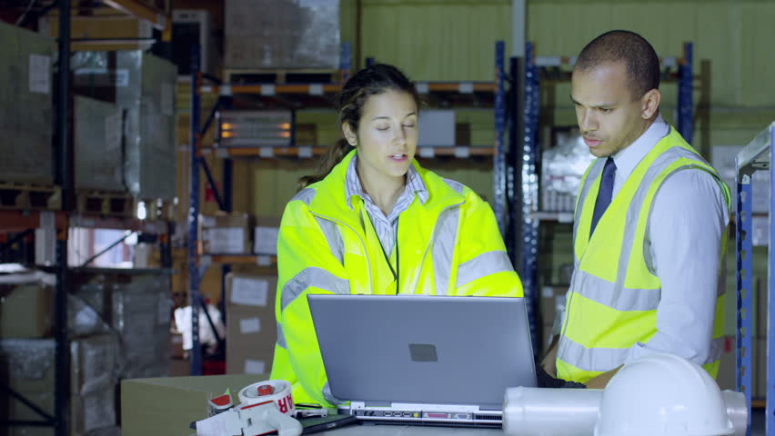 2 warehouse workers in high visibility clothing are discussing their business and looking at a laptop computer. A third worker interrupts them to ask a question.
