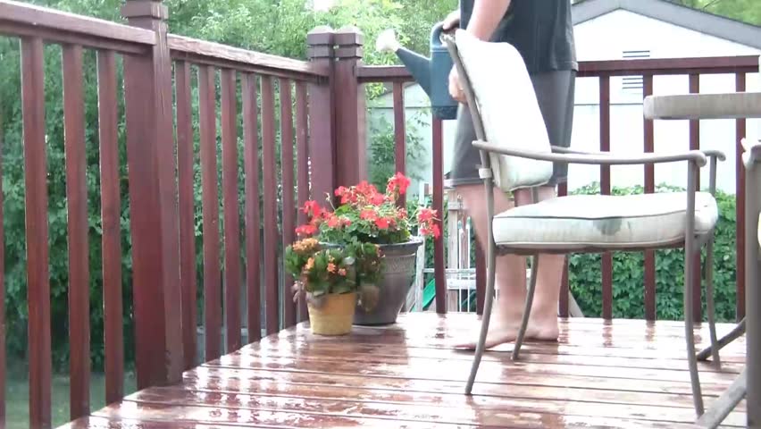 Man stands on deck in and ironically waters flowers in the rain in summer attire.