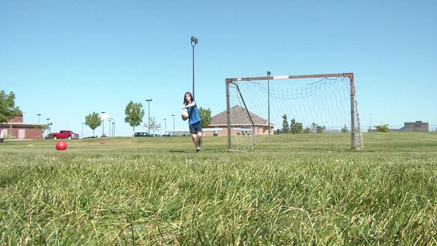 Girl stands inside goal in sunlight while foot kicks soccer ball towards her, saving one, the other bouncing off cross bar.