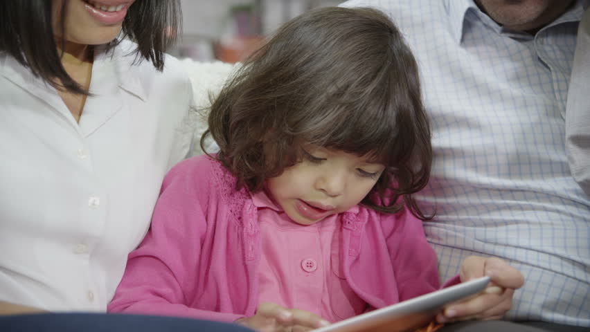 Mother and father of Asian ethnicity are bonding with their children at home. Their young daughter plays on a tablet computer and begins to giggle. Slow motion.