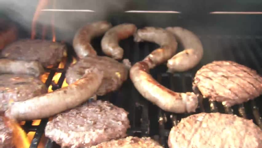 Clip of bratwurst and hamburgers being grilled over flames with smoke, in sunlight.