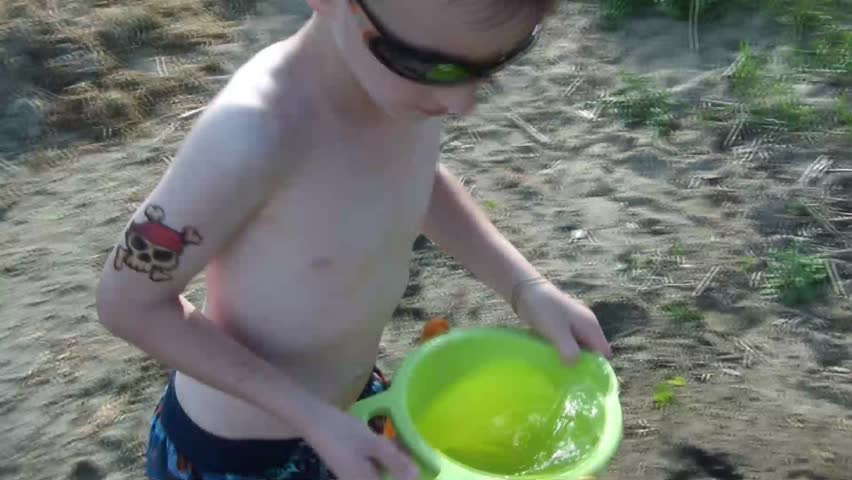 Boy plays on beach with bucket of water that he pours onto sand, looks up and smiles in sun, and then proceeds to start filling bucket with sand.