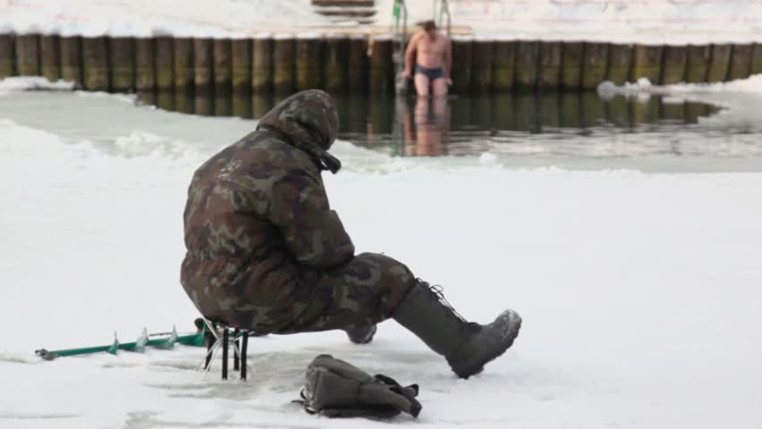 Fisherman in warm coverall sit near ice hole and man swims at winter day