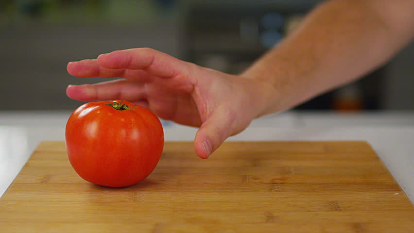 A medium dolly to static shot of two organic tomatoes being placed on a lightly-colored wooden cutting board, as they're being prepared by a chef. Filmed on a RED Epic.