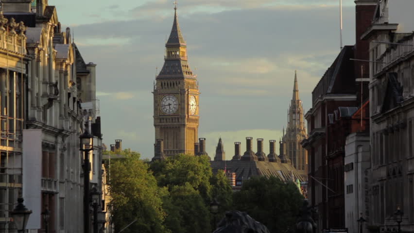 A stationary shot of Big Ben clock tower in London as seen from distance. There are houses and treetops on a street before the tower. 