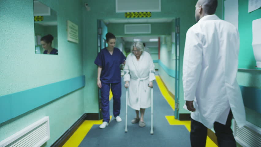 An attractive female medical assistant or nurse helps an elderly lady on crutches to take a walk down the hospital corridor. In slow motion.