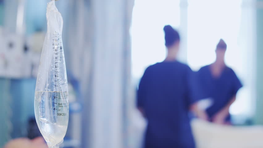 Close up of an IV drip at an elderly patients bedside. In the background two nurses can be seen attending to another patient. In slow motion.
