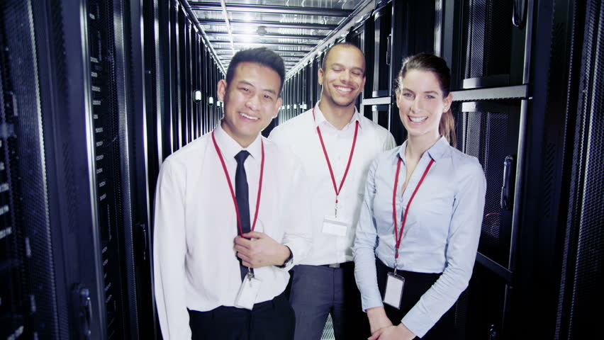 Three people of mixed ethnicity are working in a data center with rows of server racks and super computers. They are walking up and down and checking all of the equipment. 