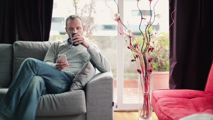 Young man drinking tea and using smartphone on sofa at home
