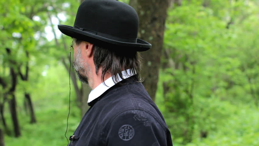 killer shoots in the spectator. Wearing a suit and hat, and old fashion eyeglasses. Outdoors.