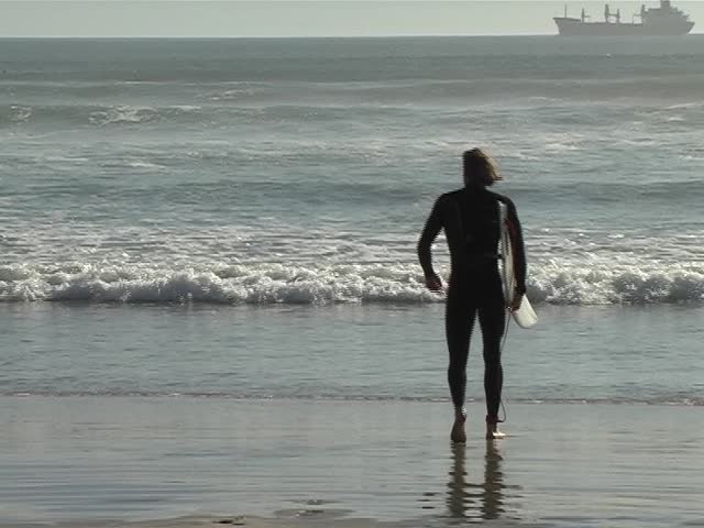 Young blonde man walking and running with his surfboard into the waves Ã¢â¬â Silhouette, with ships in background