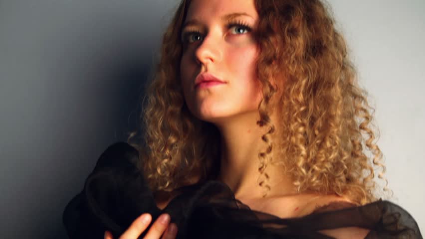 Young model with curly hair stands near wall wrapped in black shawl, shown in motion