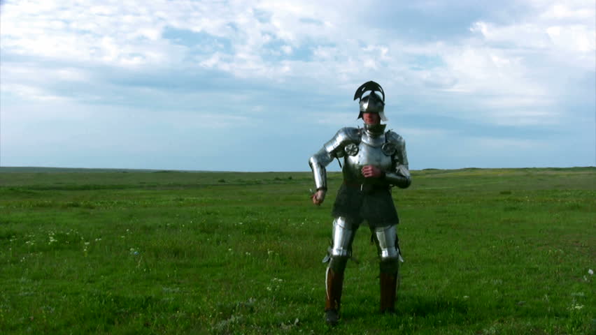 Medieval knight in armor, and with an open visor exercise with a sword against the backdrop of steppe grass and blue sky