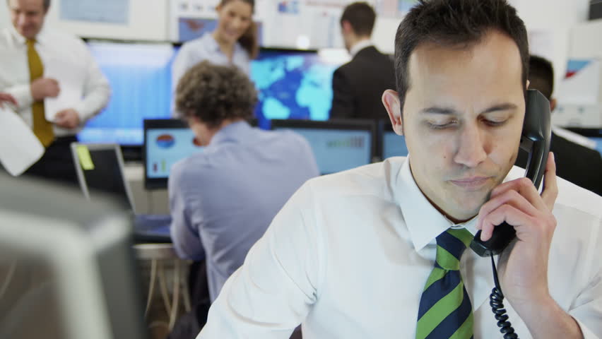 Young and ambitious stock market trader is doing a deal over the phone in a busy office filled with computers. The rest of his team are hard at work in the background. In slow motion.