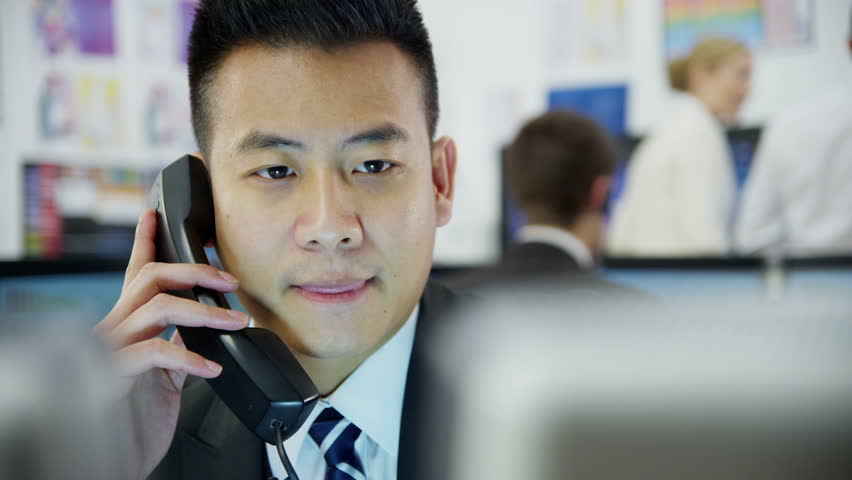 Young and ambitious stock market trader is doing a deal over the phone in a busy office filled with computers. The rest of his team are hard at work in the background. 