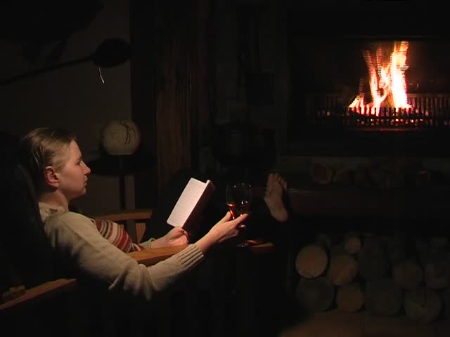 Woman sitting in front of a fire, reading and enjoying a glass of wine