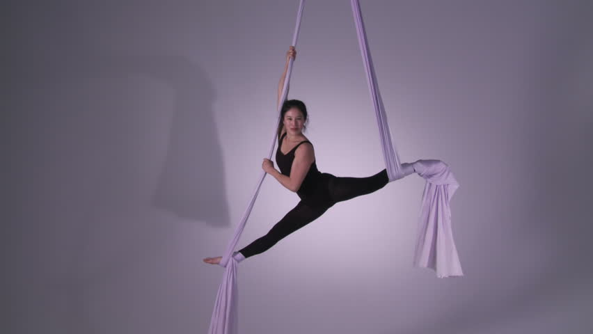 Gymnastic young woman does splits and spins while doing aerial yoga. Wide shot with lilac tint and neutral backgrounds.