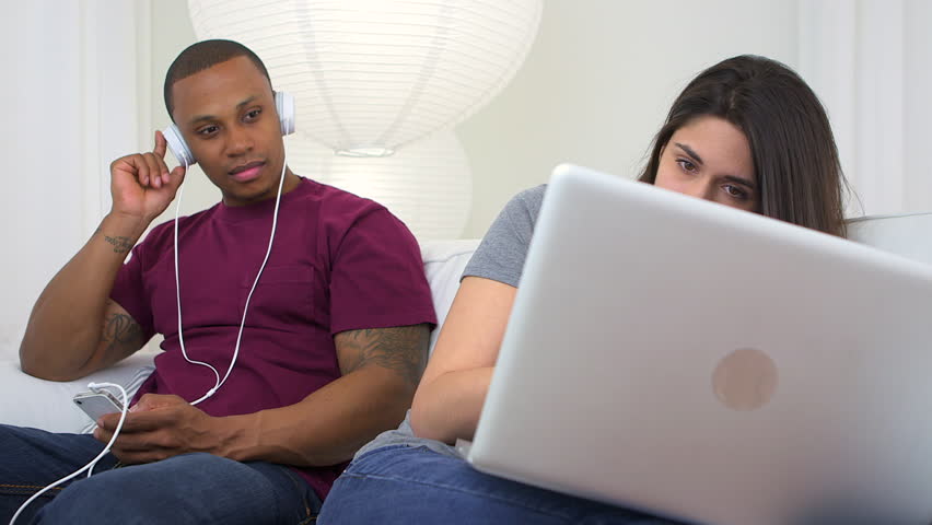 African American and Caucasian couple using earphones and computer on couch