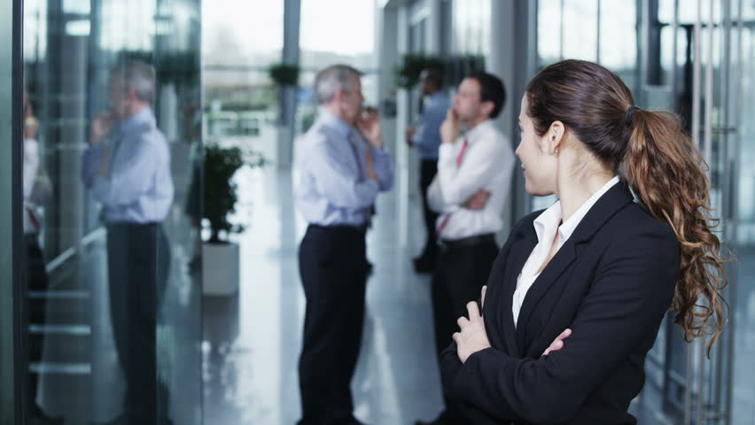 Portrait of an attractive and confident young business woman standing in a light and modern office building. The rest of her team can be seen chatting together in the background. 