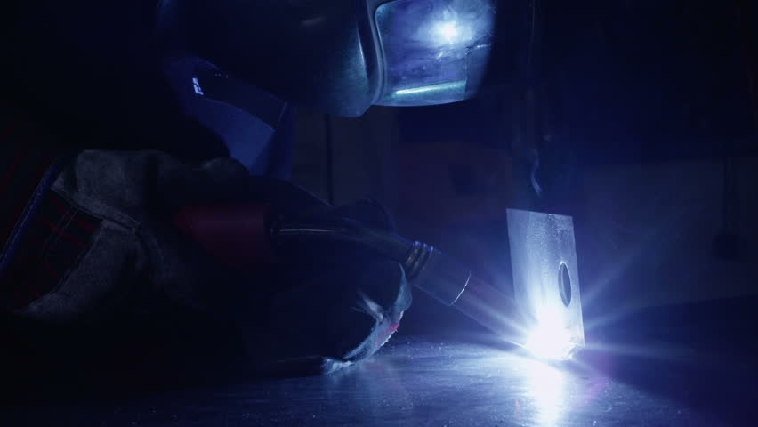 A welder is working in a dark construction or engineering plant. The flare of the flame is reflected in the visor of his mask.