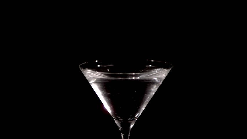 A fresh green olive on a cocktail stick is dropped in slow motion into a glass of martini, causing the drink to splash and spill. On a black background.