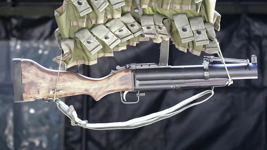 Grenade Launcher Hanging Against Black Background With Part Of Ammunition Vest Visible
