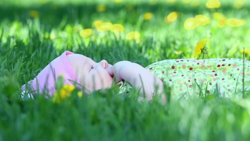 Baby girl lies and suck her finger on grass with dandelions at summer day