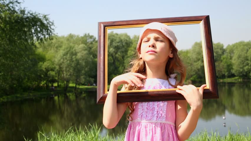 Little girl puts picture frame on head and sways, then she approaches closer, near pond at summer park