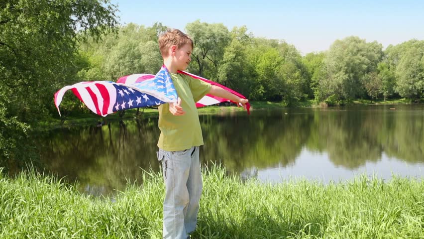 Boy in green t-shirt holds unfolded US flag which flutters on wind near pond at sunny summer day