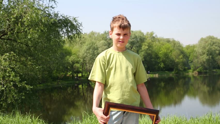 Young boy raises picture frame and approaches close, on bank of pond in park at summer sunny day