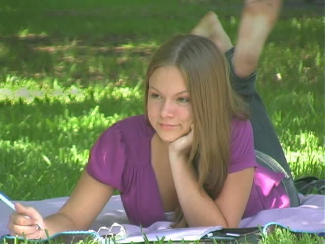 A beautiful young girl relaxing in the park, lying on a blanket and writing - doing homework, perhaps, or writing a letter.  Maybe writing in her journal.  She pauses to look at the camera and smile.