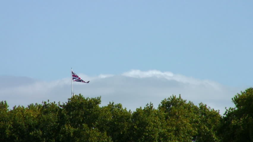 Big wide shot of Union Jack flag fluttering in a light summer breeze. Foreground green park trees and background sky and clouds. Tripod locked-off .HD1080i60