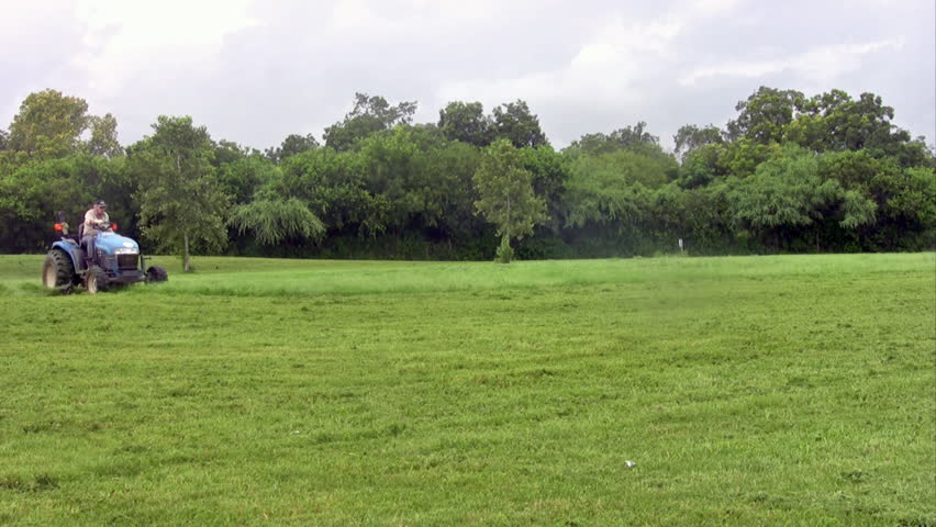 Video of large city owned tractor lawn mowers cutting grass in park. Tractors turning corner. Rain and drizzle, wet grass being cut. Green healthy grass and forest in city park. High speed tractors.