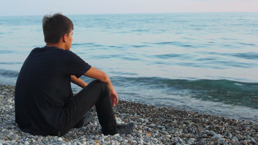 teenager sits on pebble beach and looks at sea, back view 