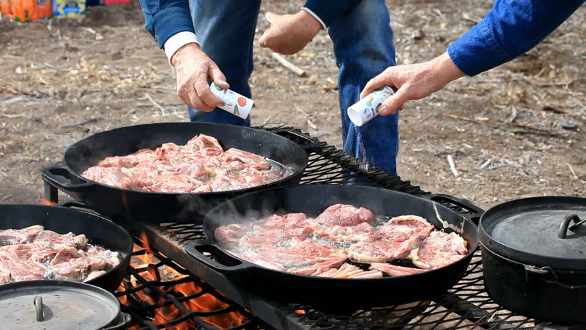 Lamb steaks and chops being salt and pepper while cooking in caste iron pans over an open outdoor fire. Cowboy and sheep camp bbq and party. Fresh meat with smoke and oil boiling near Dutch Oven.
