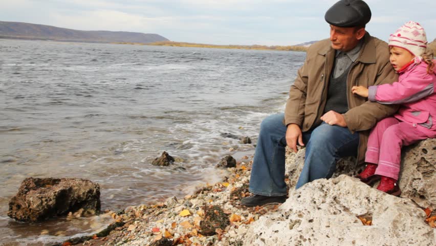 daddy with daughter sit on stone on river bank and look at waves 