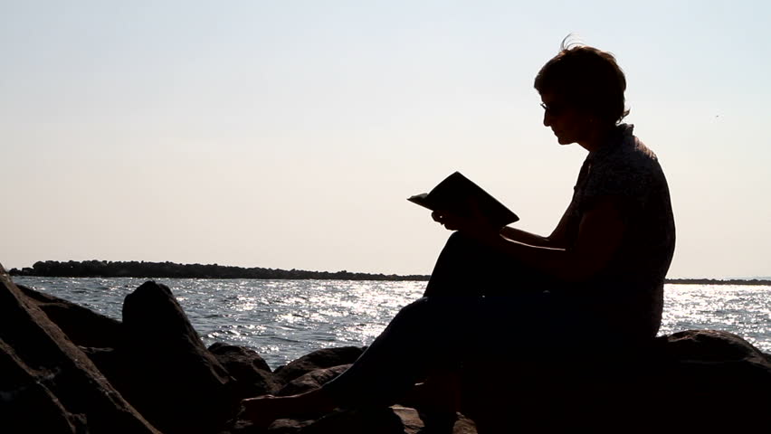 Silhouette of woman sitting on a rock studying the bible by a body of water.