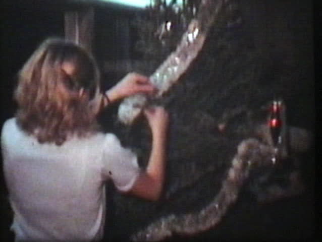 Three teenage siblings begin decorating the Christmas tree by putting the silver garland around the tree.