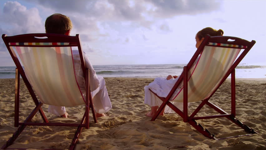 An elderly couple sits on the beach together and holds hands.